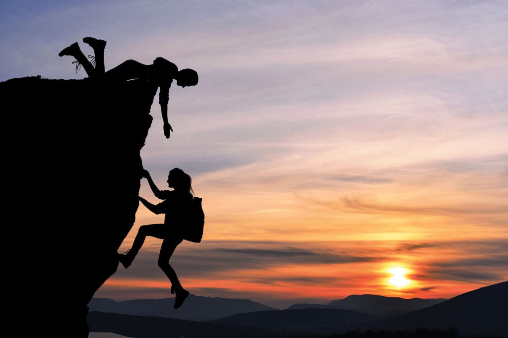 At sunset, one climber lies at the top of a cliff and reaches down to help another climber up the rock face